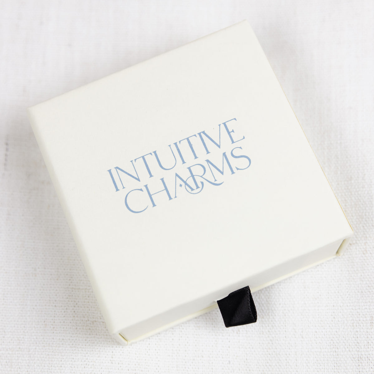 A cream-colored square jewellery gift box with the words 'INTUITIVE CHARMS' printed in light blue on the lid, resting on a white textured fabric background.