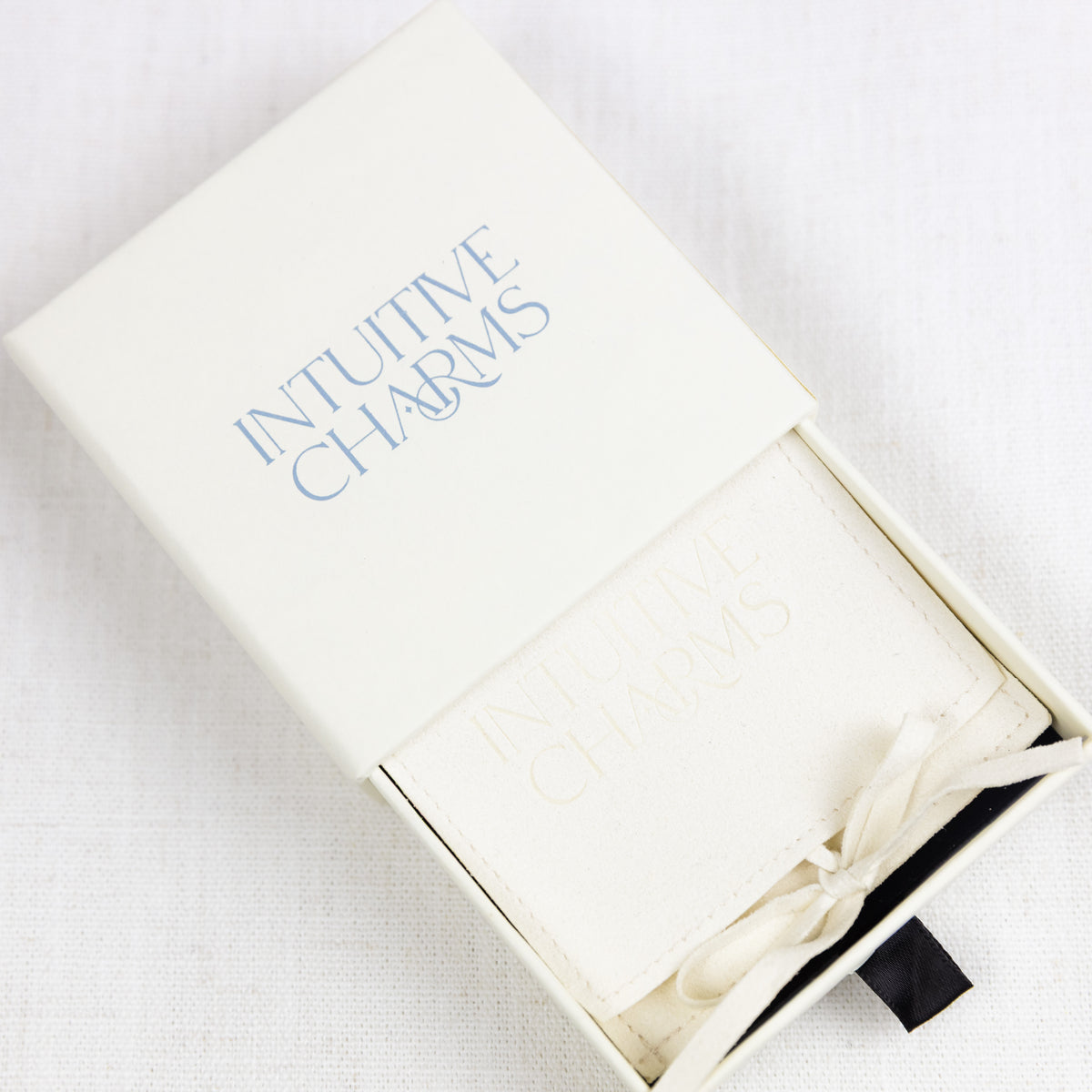 An off-white leatherette Intuitive Charms jewellery pouch inside its matching cream-colored presentation box, which features the brand name 'INTUTITIVE CHARMS' in light blue text on the lid.