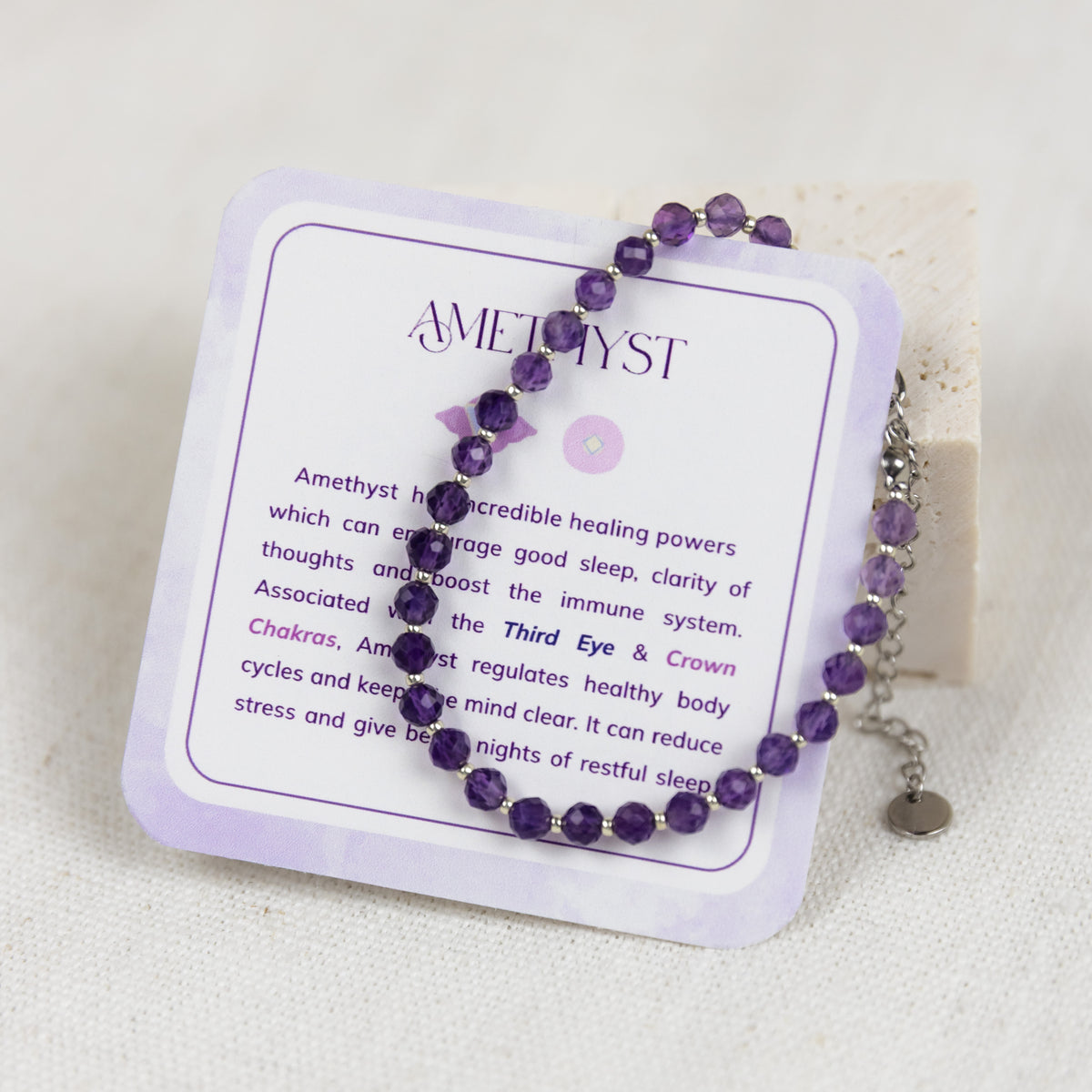 An amethyst beaded bracelet from intuitive charms draped over a white display card with purple text, set against a soft beige fabric background.