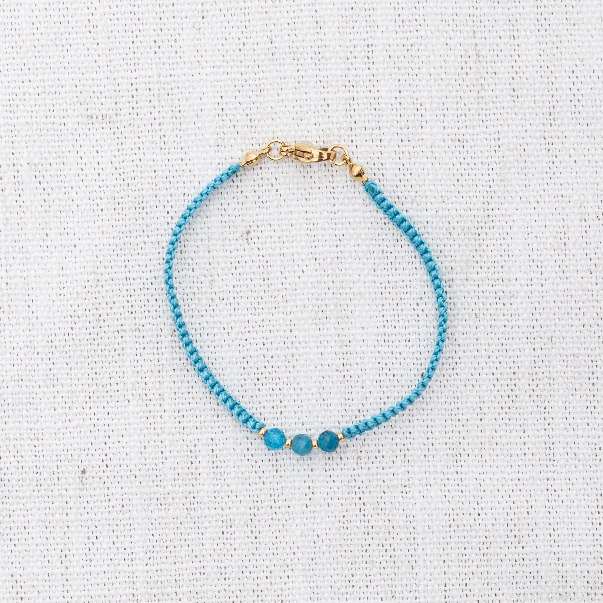 Intuitive Charms Delphina's Guard Apatite Braided natural Crystal beaded custom Bracelet in gold and blue cord and clasp on a light fabric background