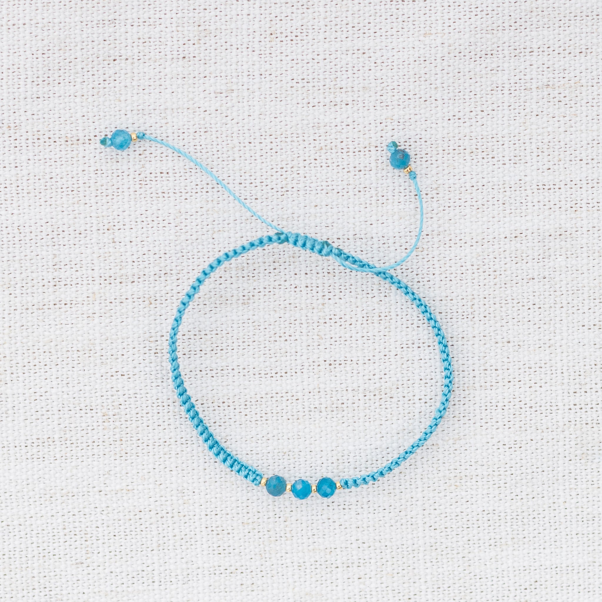 Intuitive Charms Delphina's Guard Apatite Braided natural Crystal beaded custom Bracelet in gold and blue cord on a light fabric background