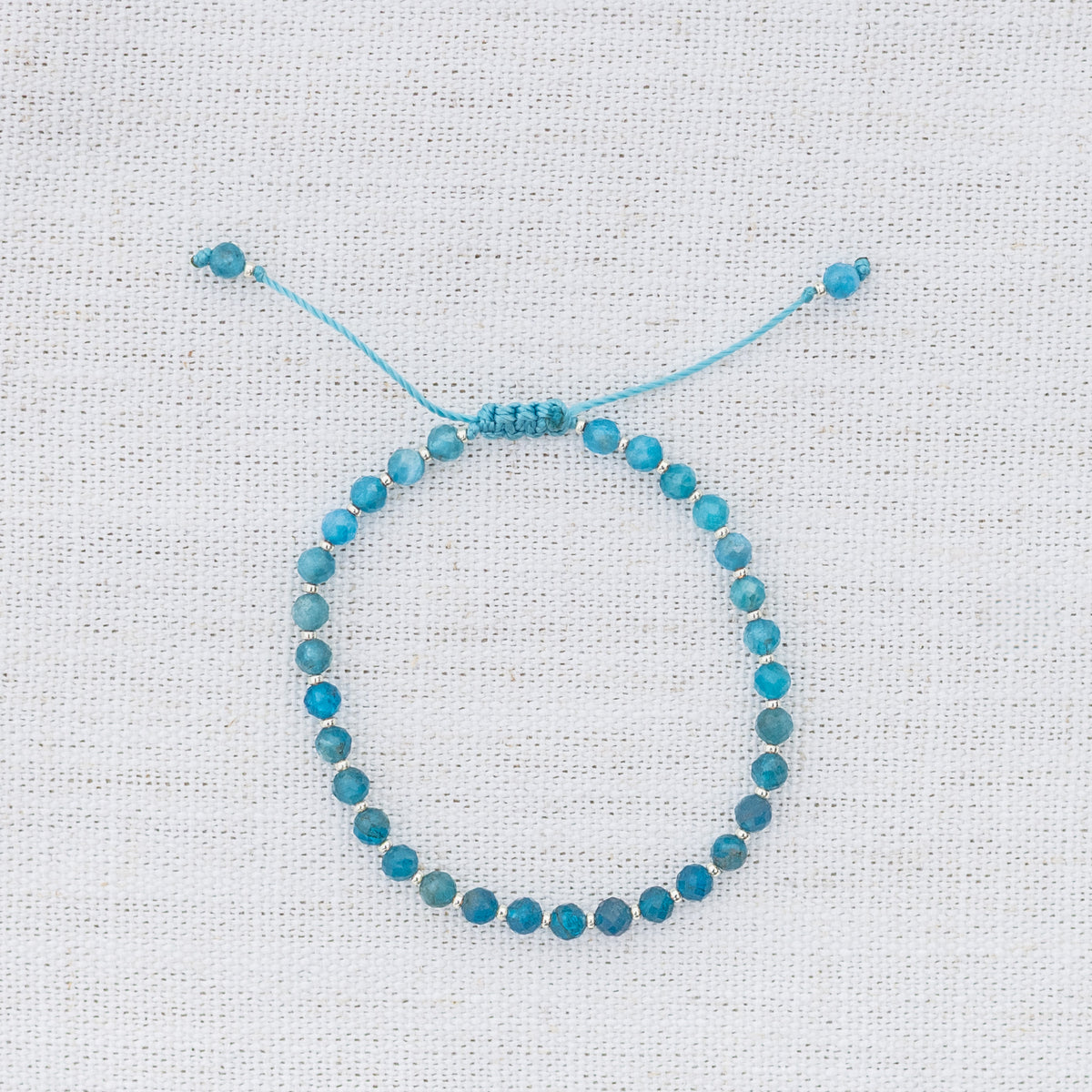 Intuitive Charms Delphina's Glow Apatite Braided natural Crystal beaded custom Bracelet in silver and blue cord on a light fabric background