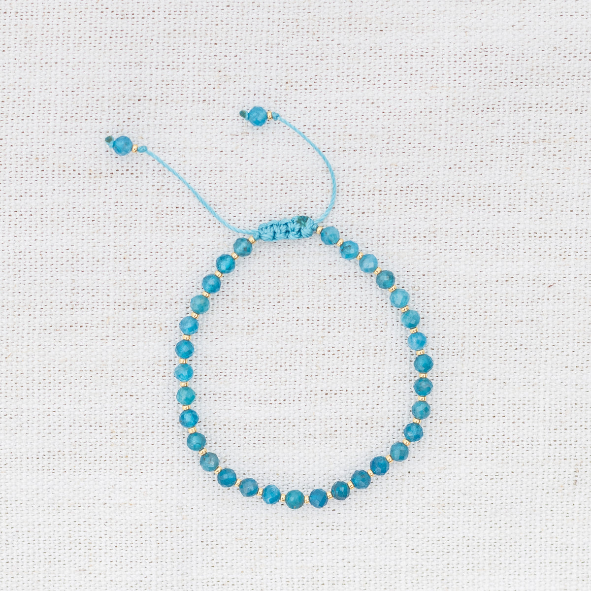 Intuitive Charms Delphina's Glow Apatite Braided natural Crystal beaded custom Bracelet in gold and blue cord on a light fabric background