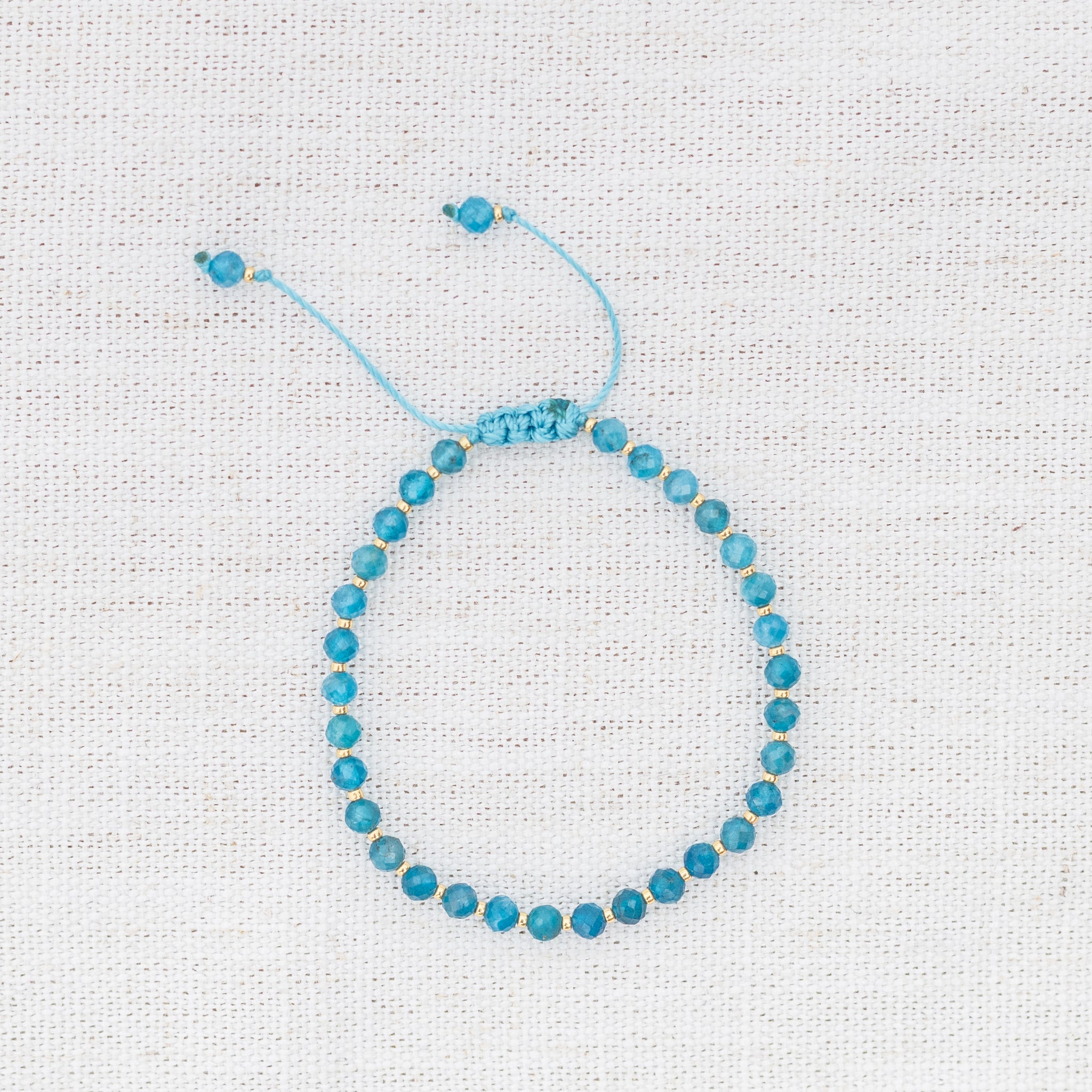 Intuitive Charms Delphina's Glow Apatite Braided natural Crystal beaded custom Bracelet in gold and blue cord on a light fabric background