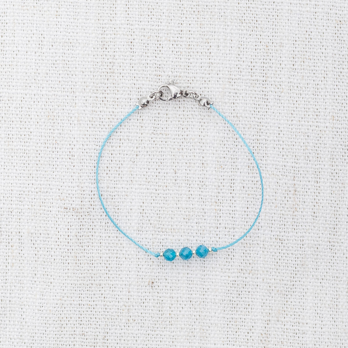 Intuitive Charms Delphina's Mist Apatite Braided natural Crystal beaded custom Bracelet in silver and blue cord and clasp on a light fabric background