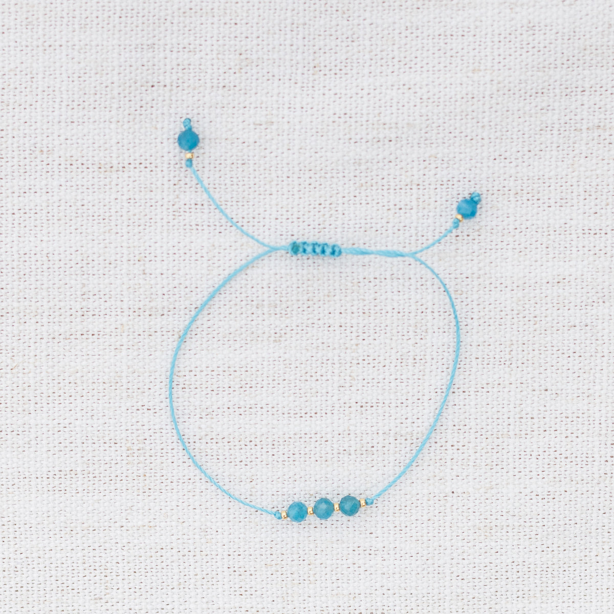 Intuitive Charms Delphina's Mist Apatite Braided natural Crystal beaded custom Bracelet in gold and blue cord on a light fabric background
