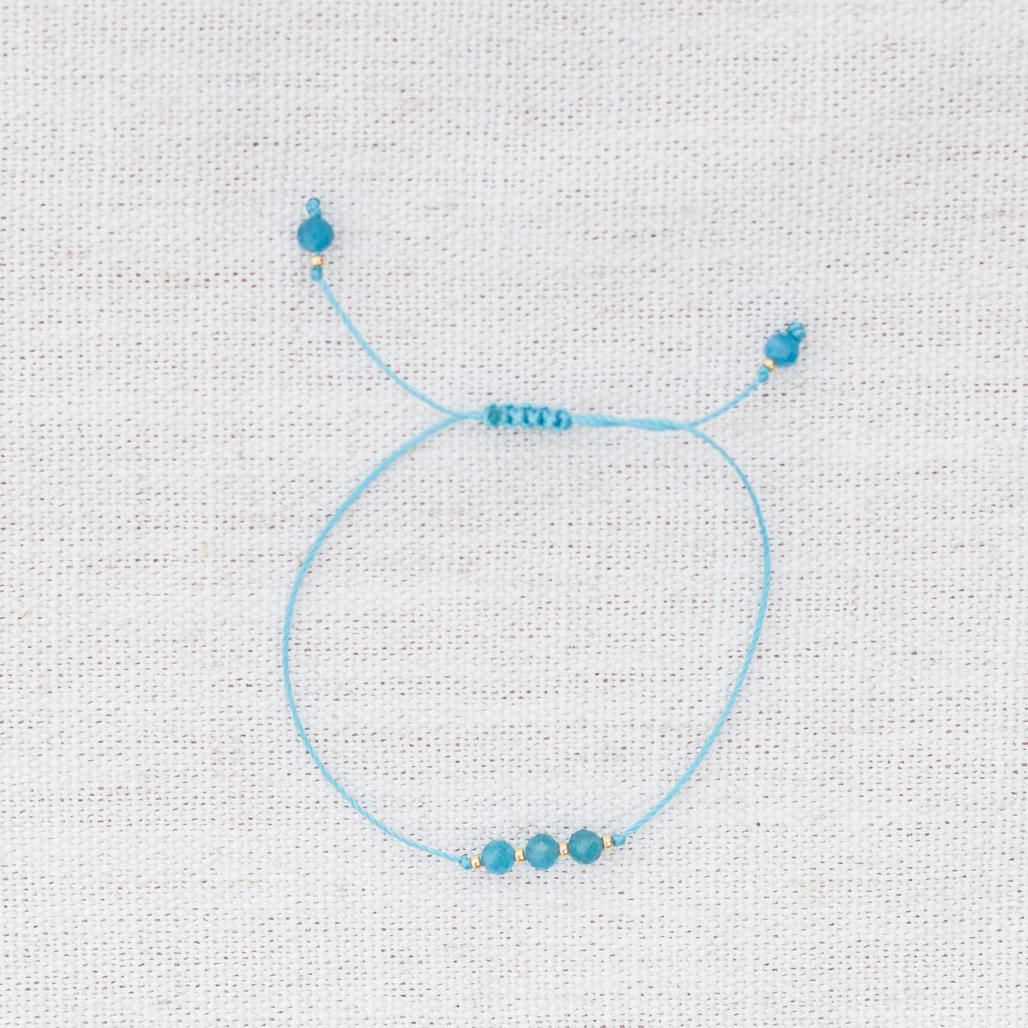 Intuitive Charms Delphina's Mist Apatite Braided natural Crystal beaded custom Bracelet in gold and blue cord on a light fabric background