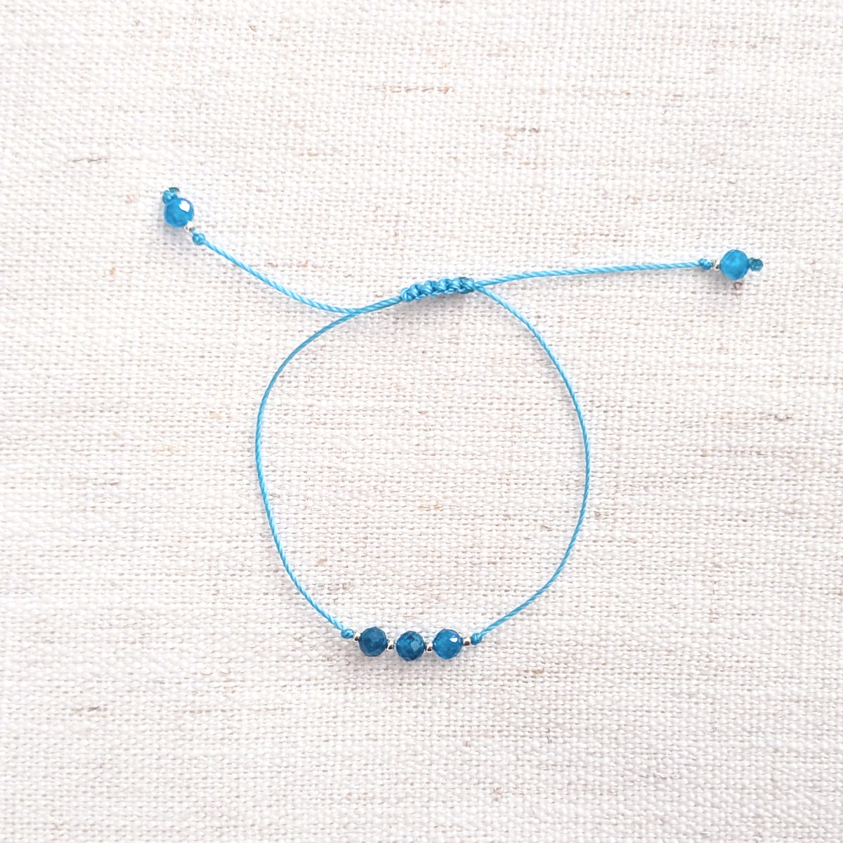 Intuitive Charms Delphina's Mist Apatite Braided natural Crystal beaded custom Bracelet in silver and blue cord on a light fabric background