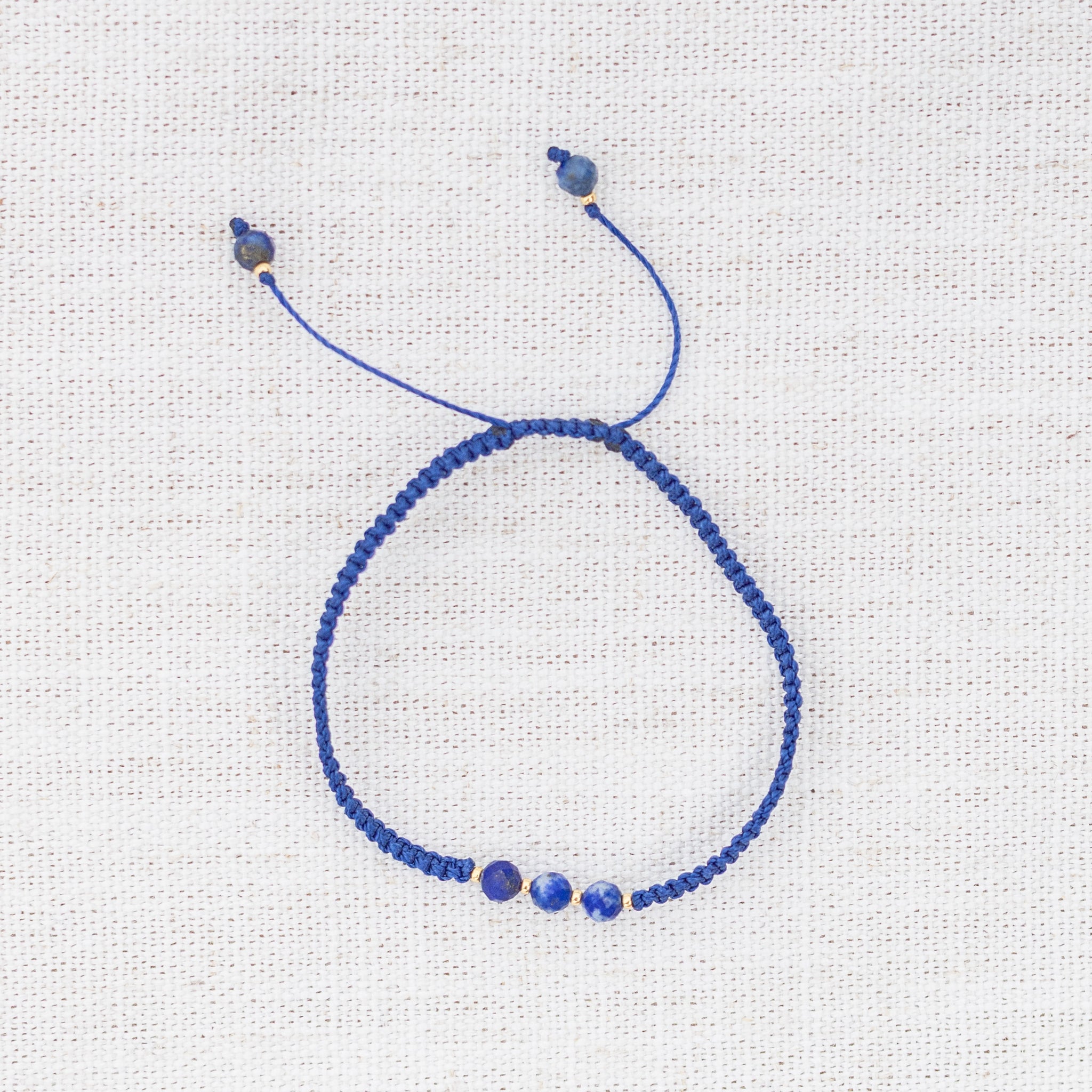 Intuitive Charms Amphitrite's Guard Lapis Lazuli Braided natural Crystal beaded custom Bracelet in gold and deep blue cord on a light fabric background