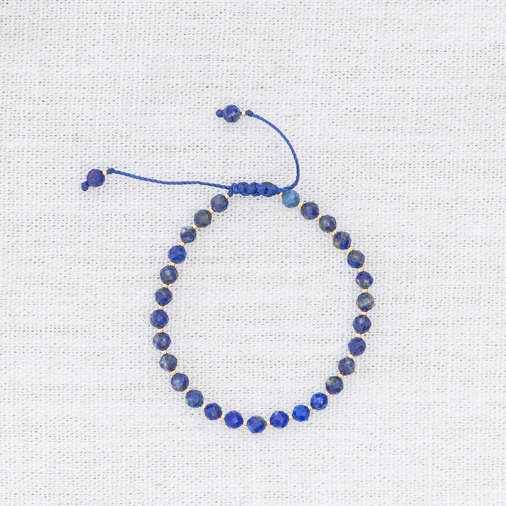 Intuitive Charms Amphitrite's Glow Lapis Lazuli Braided natural Crystal beaded custom Bracelet in gold and deep blue cord on a light fabric background