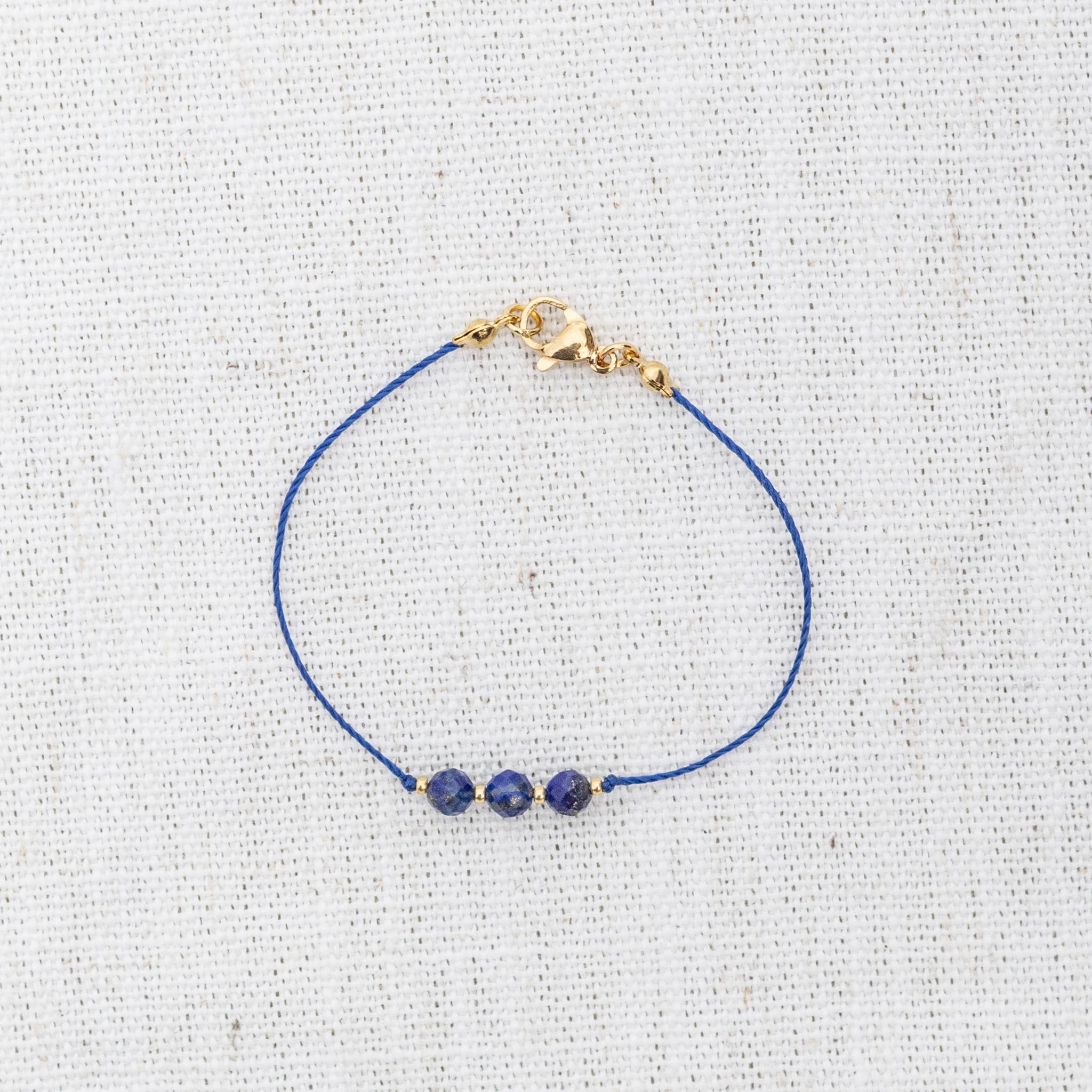 Intuitive Charms Amphitrite's Mist Lapis Lazuli Braided natural Crystal beaded custom Bracelet in gold and deep blue cord and clasp on a light fabric background