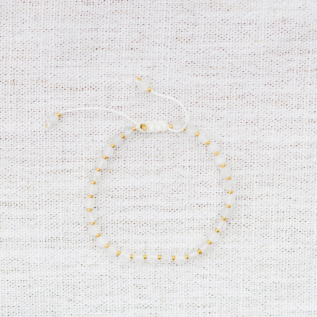 Intuitive Charms Selene's Glow Moonstone Braided natural Crystal beaded custom Bracelet in gold and white cord on a light fabric background