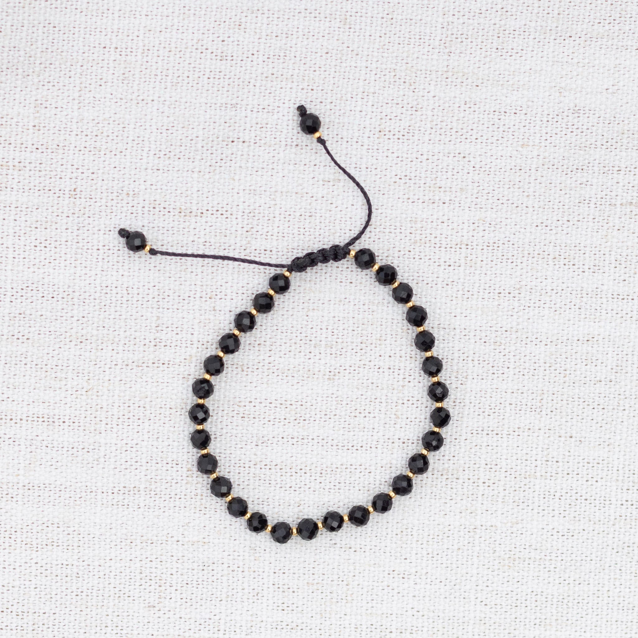 Intuitive Charms Ceto's Glow Apatite Braided natural Crystal beaded custom Bracelet in gold and black cord on a light fabric background