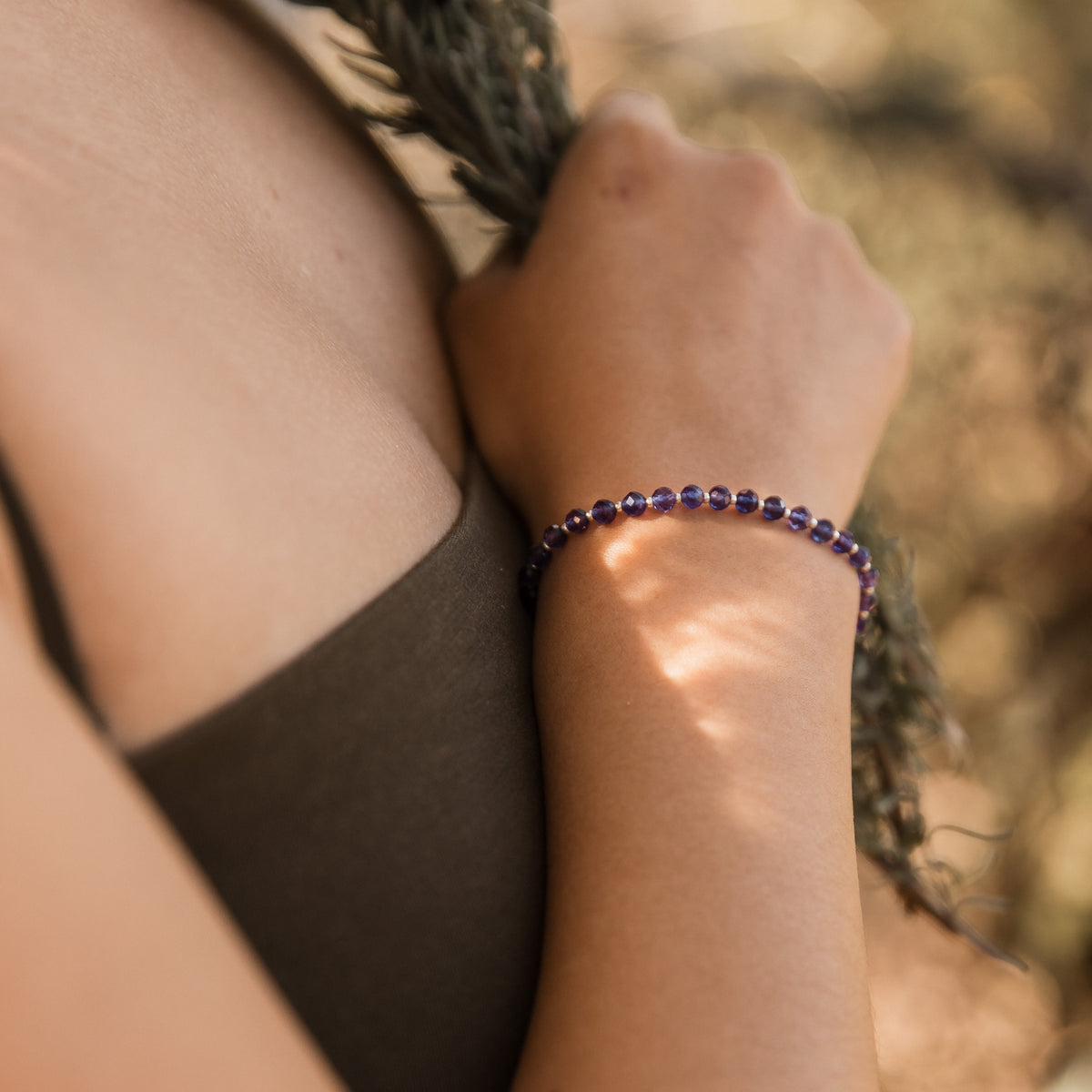 A close-up lifestyle shot of a person wearing a beaded bracelet on their wrist, holding a dark feather. The bracelet consists of small, dark purple amethyst faceted beads separated by tiny silver spacer beads. Made by intuitive charms.