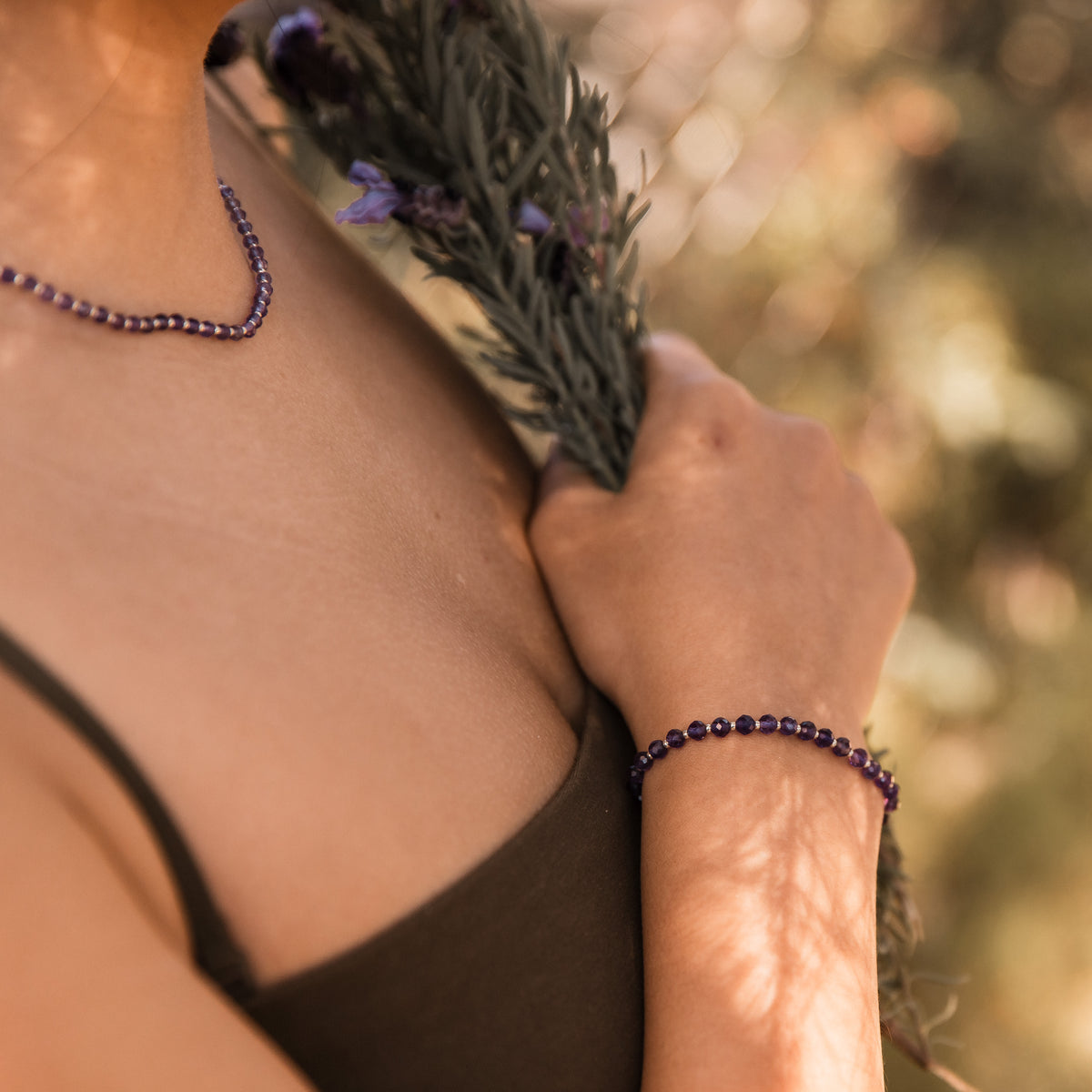 A close-up lifestyle shot of a person wearing a dark purple amethyst faceted beaded bracelet on their wrist and a matching beaded necklace on their neck, holding a sprig of lavender against a warm, blurred outdoor background. Made by intuitive charms.