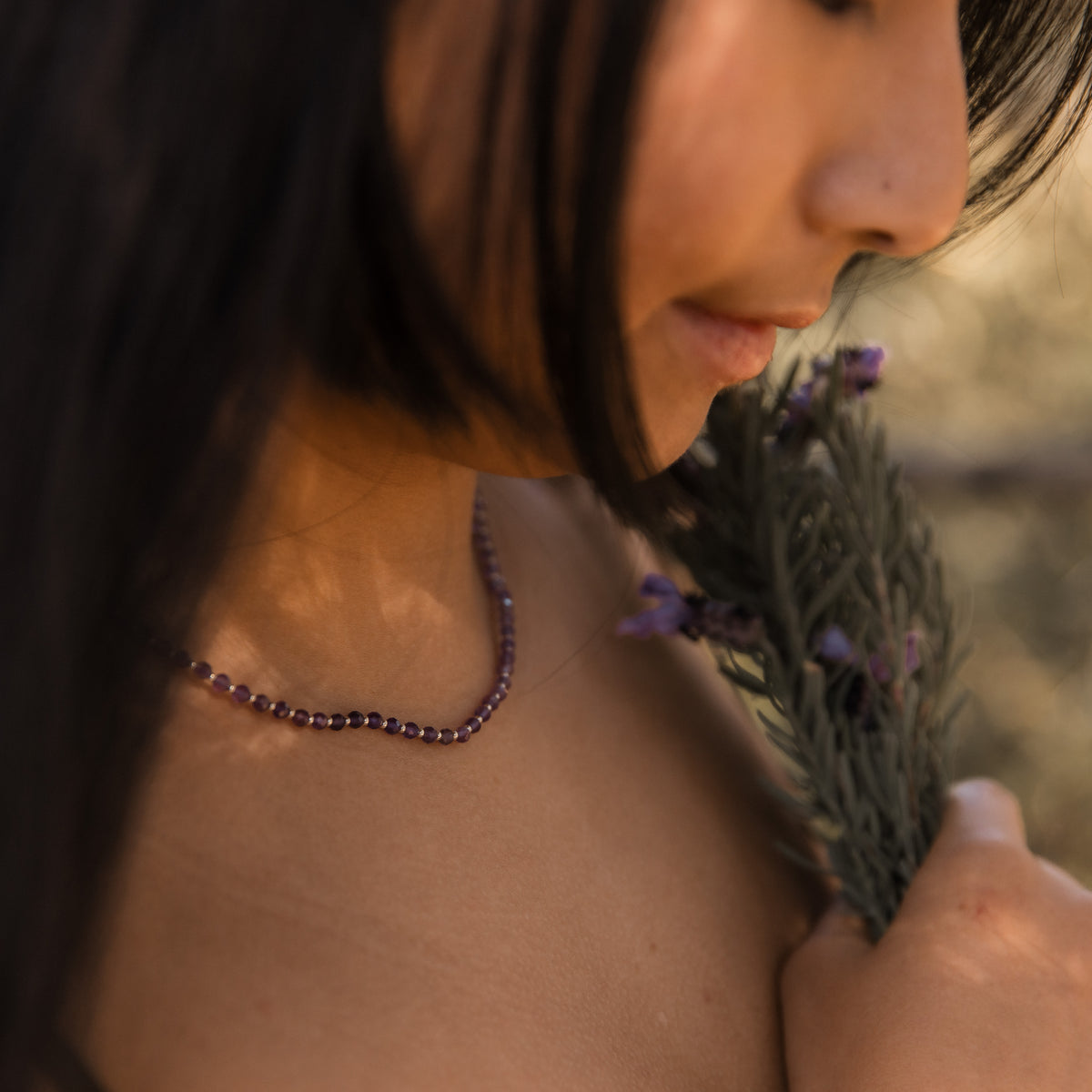 A close-up lifestyle shot of a woman wearing a dark purple amethyst faceted beaded necklace, holding a sprig of dried lavender against a warm, blurred outdoor background. Made by intuitive charms.