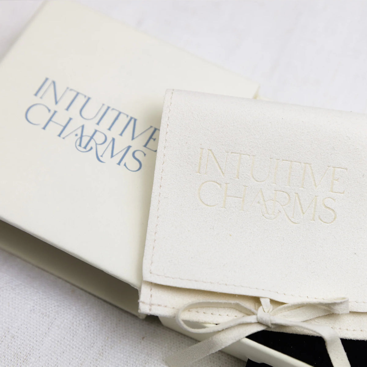 An off-white leatherette jewellery cover with 'INTUITIVE CHARMS' embossed on the flap, tied with a matching ribbon next to its matching cream-colored box.