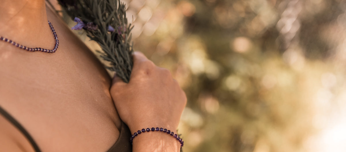 A close-up lifestyle shot of a person outdoors wearing a dark purple amethyst faceted beaded bracelet on their wrist and a matching beaded necklace on their neck, holding a sprig of lavender to their chest. Made by intuitive charms.