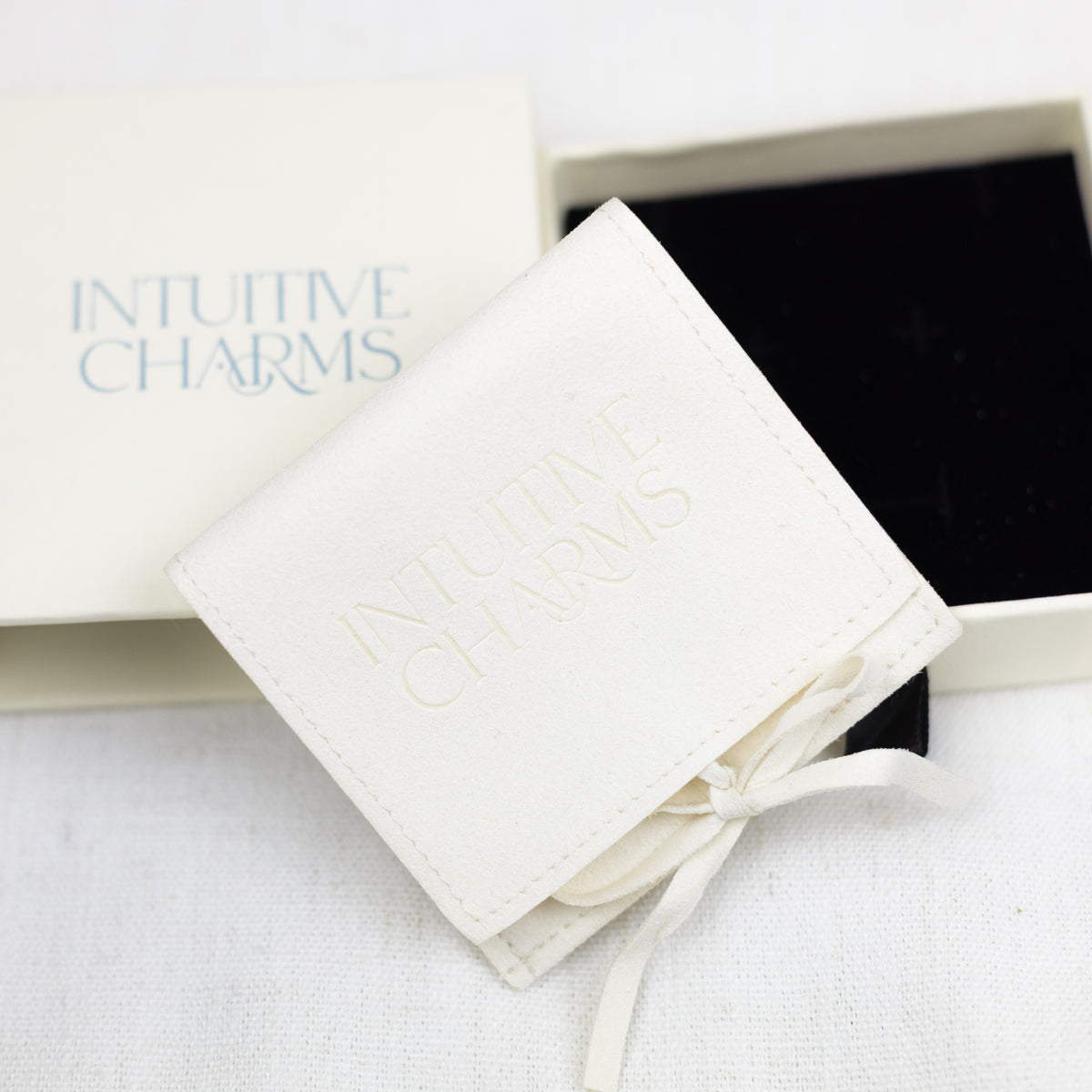 An off-white leatherette jewellery holder pouch with 'INTUITIVE CHARMS' embossed on the front, tied with a matching ribbon, displayed next to its branded cream-colored box and black insert.