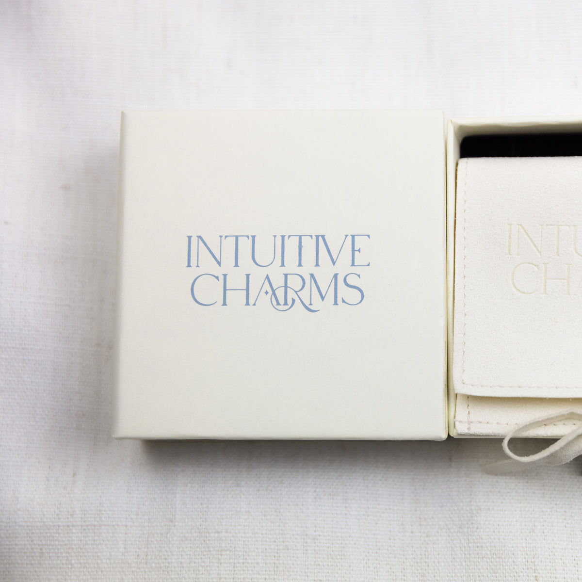 An off-white jewelry box with the brand name 'INTUITIVE CHARMS' printed in light blue on the lid, shown next to a matching off-white pouch on a textured white fabric background.
