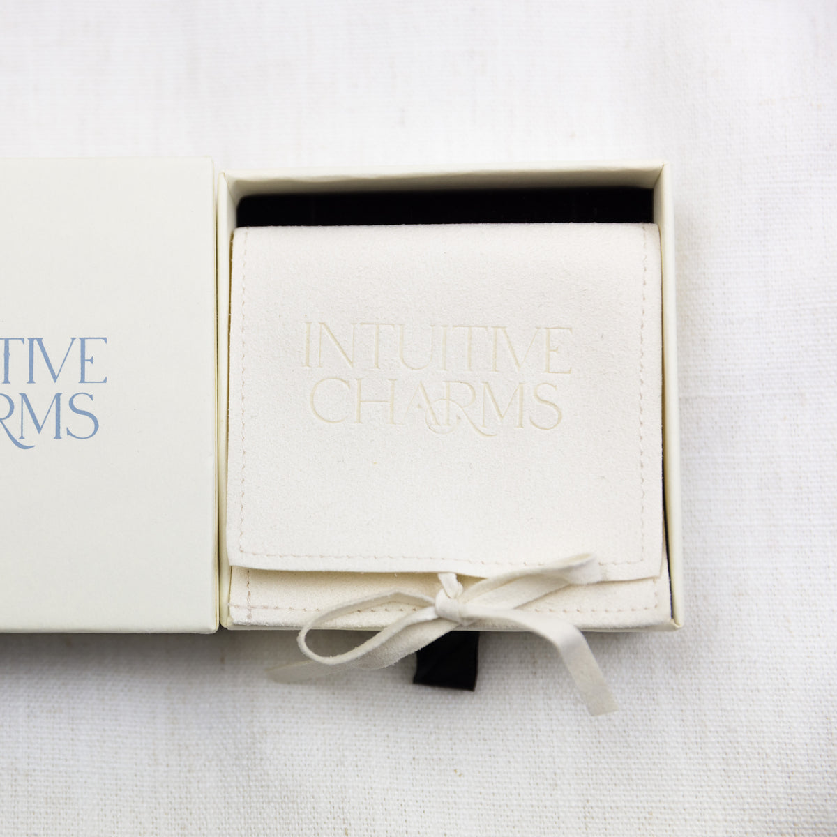 An open cream-colored presentation jewellery box on a white textured fabric background, containing a folded cream fabric pouch embossed with the words 'INTUITIVE CHARMS' in a matching cream tone. Part of the outer box lid is visible on the left, showing blue text that reads 'INTUITIVE CHARMS'.