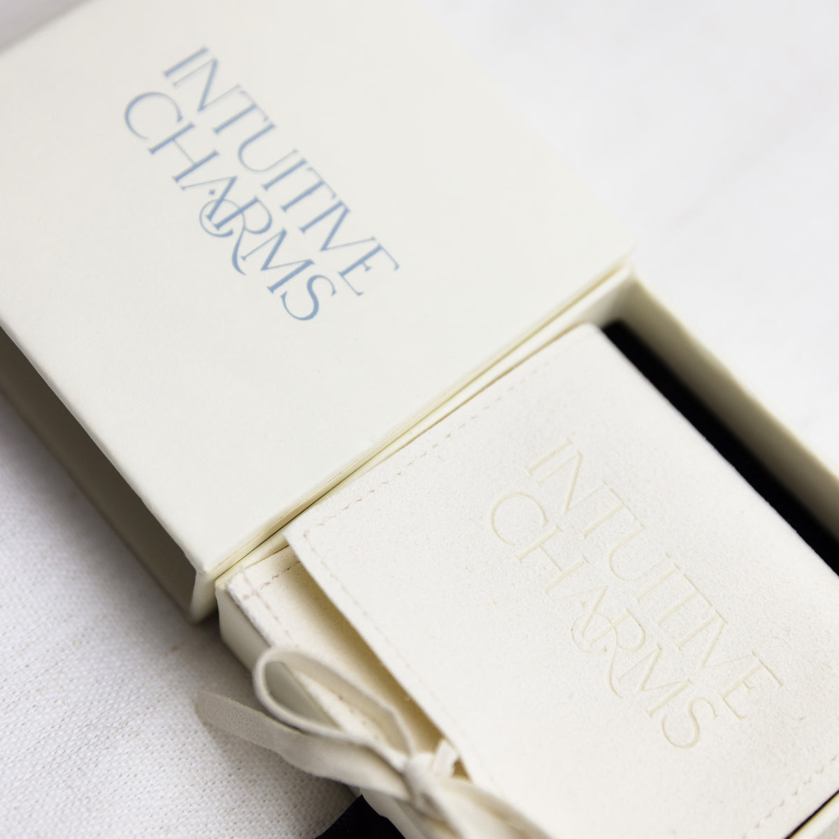 An open cream-colored presentation jewellery box with a matching cream fabric-covered card or booklet inside, both embossed with the words 'INTUITIVE CHARMS' in blue and gold tones, photographed on a light fabric surface.