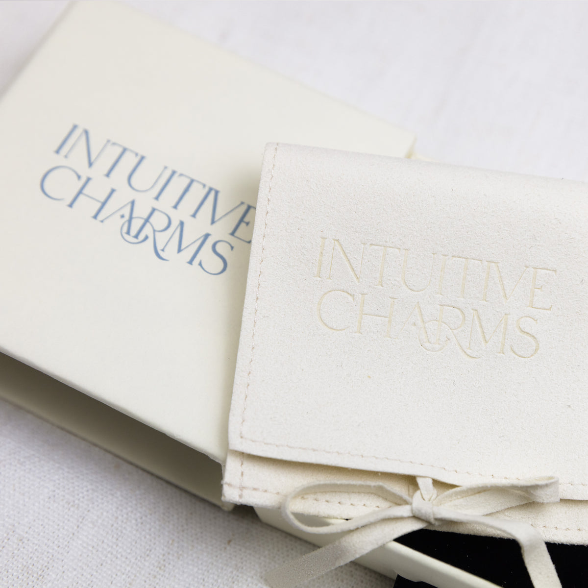 An off-white leatherette jewellery cover with 'INTUITIVE CHARMS' embossed on the flap, tied with a matching ribbon next to its matching cream-colored box.