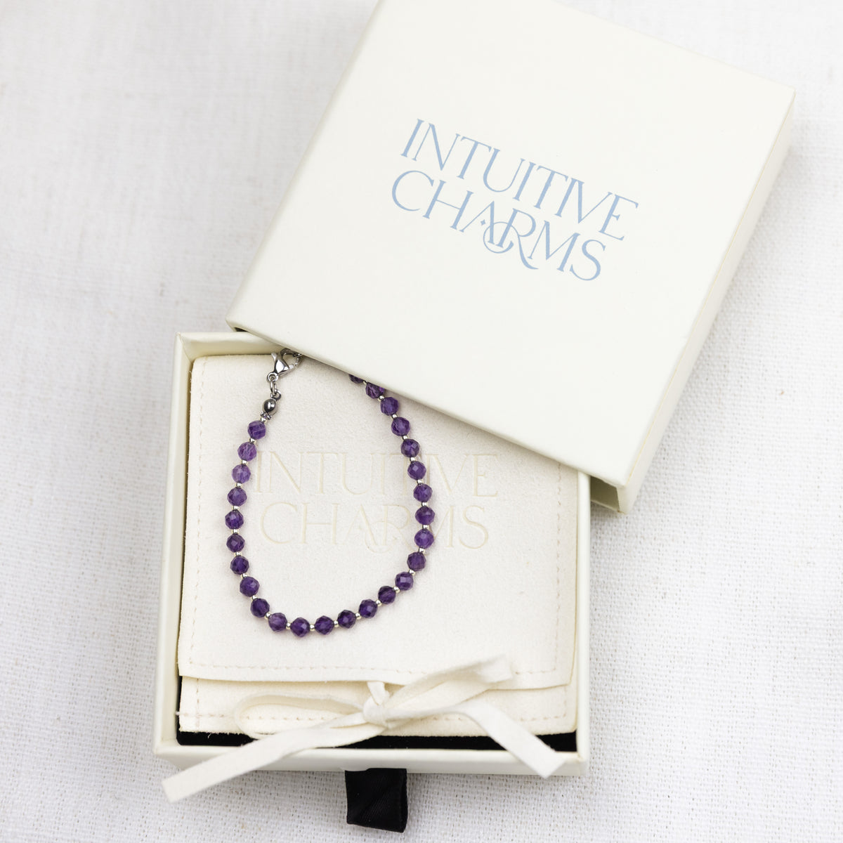 A beaded purple charm bracelet with a silver clasp, presented inside an off-white gift box with the brand name 'INTUITIVE CHARMS' printed on the lid and box interior. The box is tied with a white ribbon and set against a textured white fabric background.