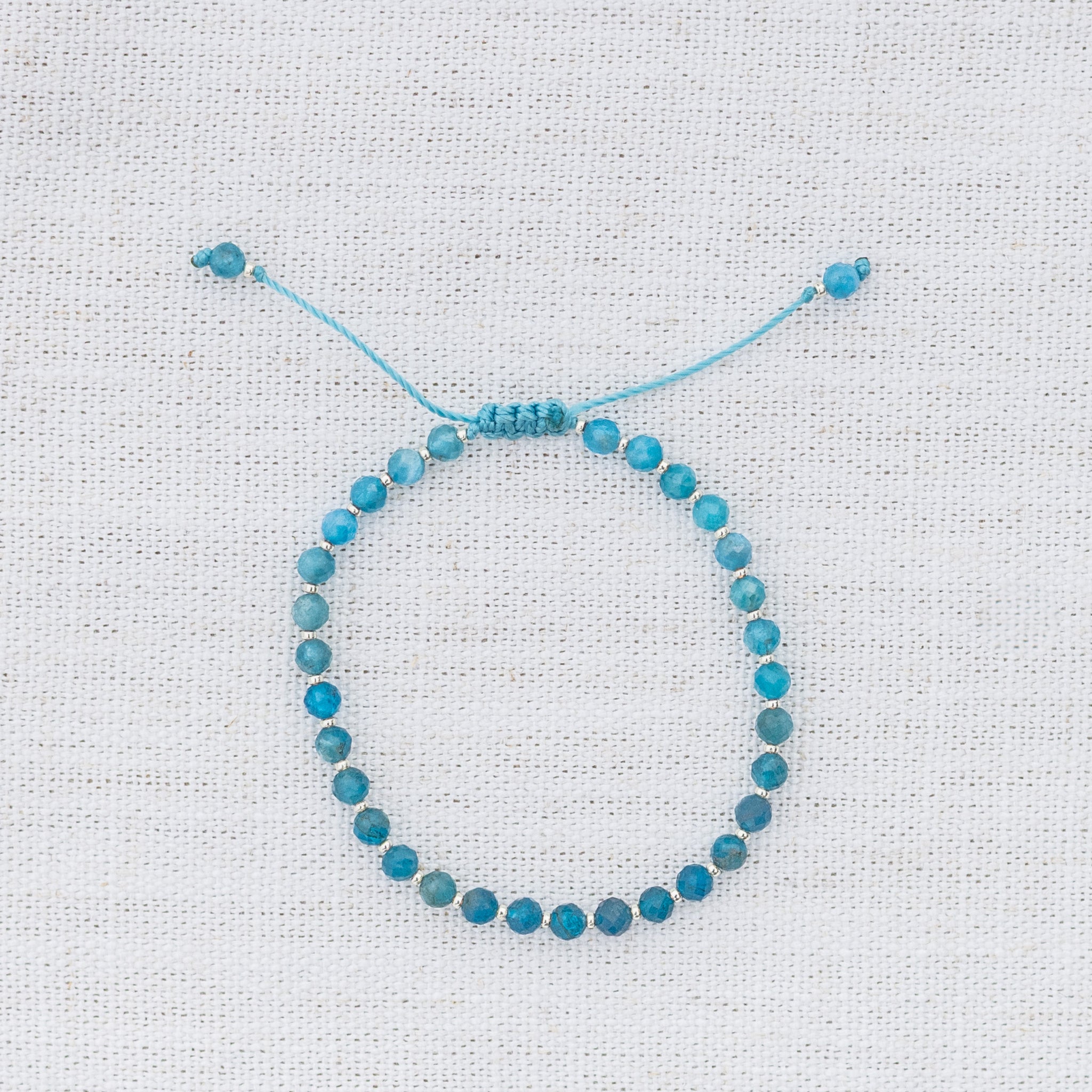 Intuitive Charms Delphina's Glow Apatite Braided natural Crystal beaded custom Bracelet in silver and blue cord on a light fabric background
