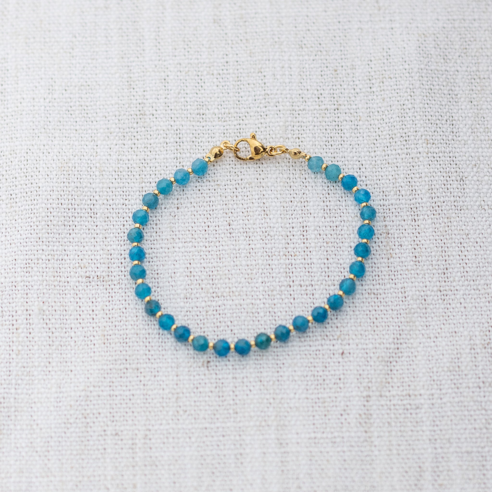 Intuitive Charms Delphina's Glow Apatite Braided natural Crystal beaded custom Bracelet in gold and blue cord and clasp on a light fabric background