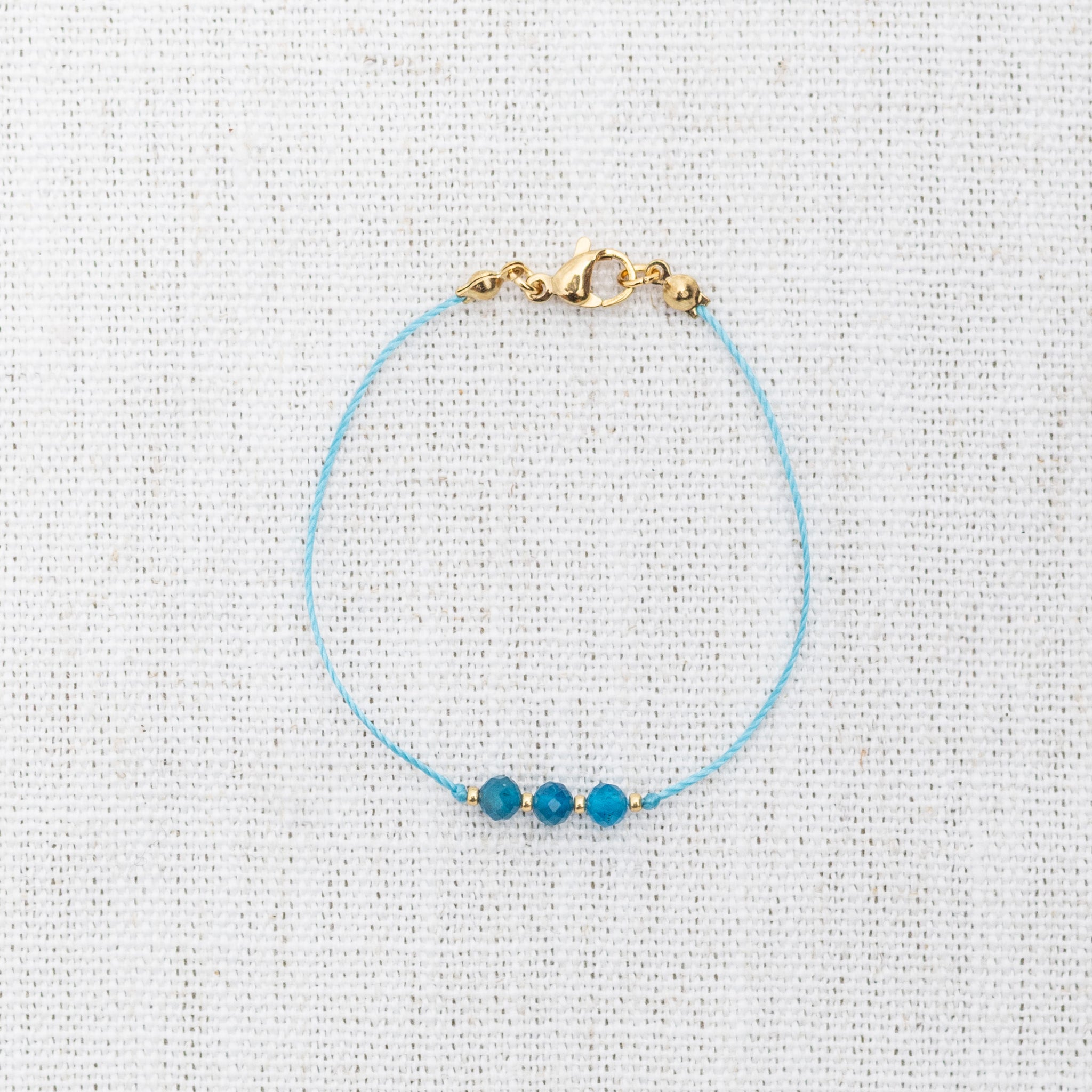 Intuitive Charms Delphina's Mist Apatite Braided natural Crystal beaded custom Bracelet in gold and blue cord and clasp on a light fabric background
