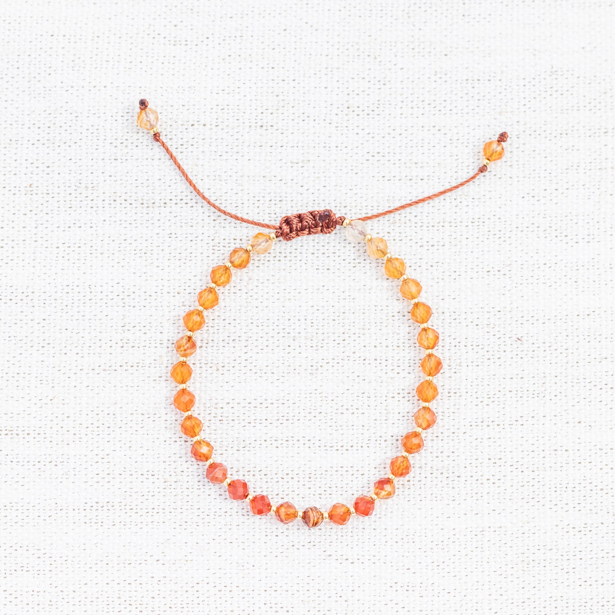 Intuitive Charms Coralia's Glow Carnelian Braided natural Crystal beaded custom Bracelet in gold and red cord on a light fabric background