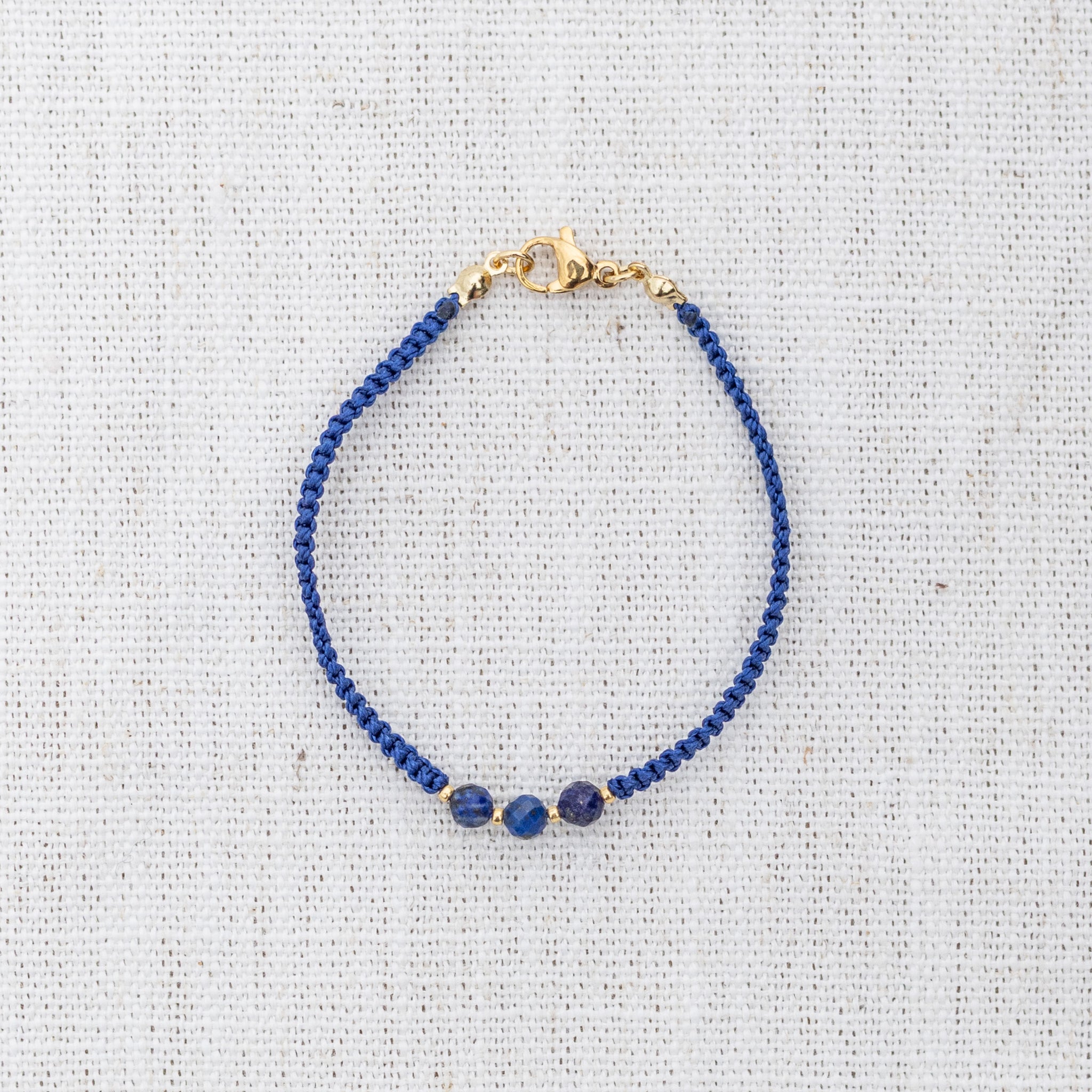 Intuitive Charms Amphitrite's Guard Lapis Lazuli Braided natural Crystal beaded custom Bracelet in gold and deep blue cord and clasp on a light fabric background
