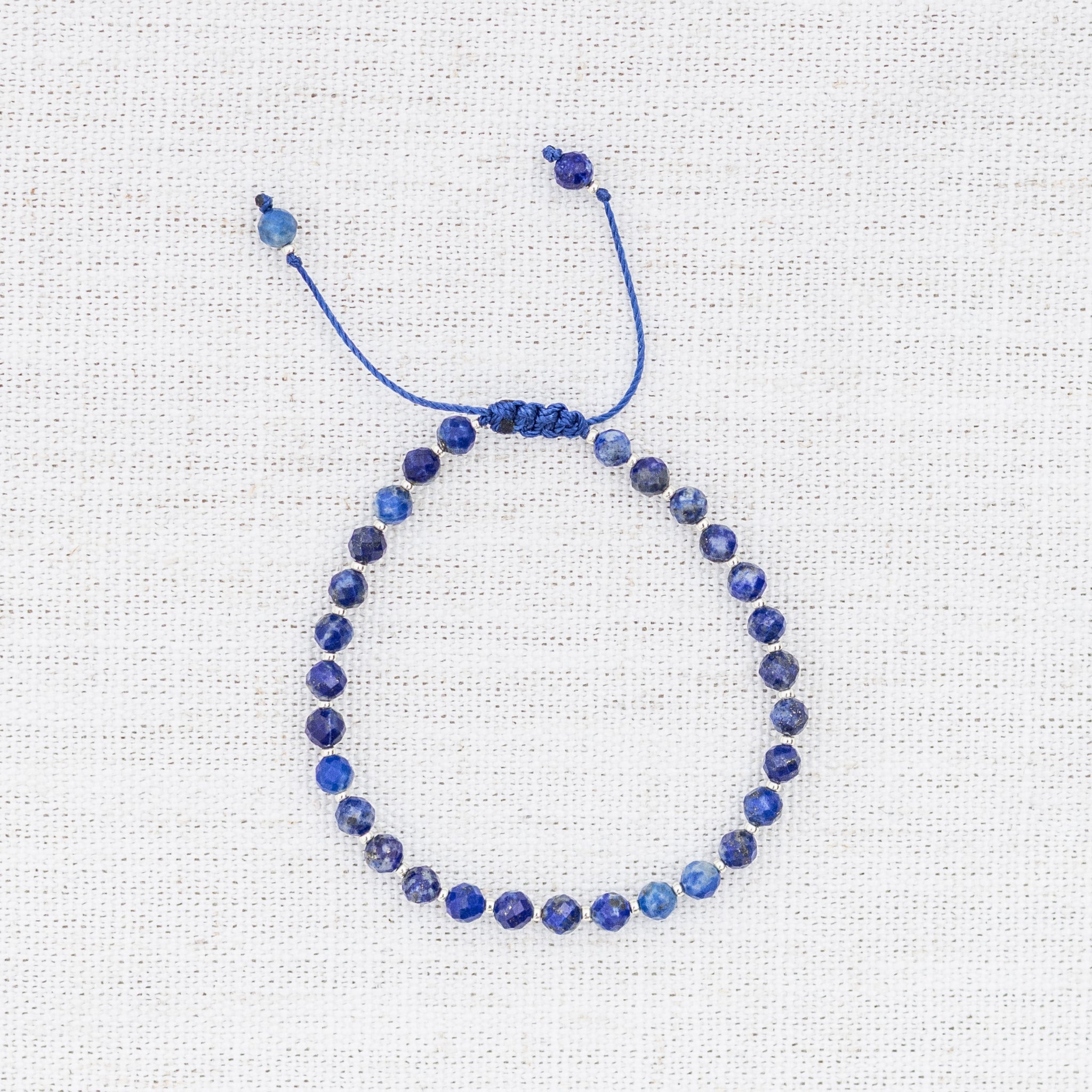 Intuitive Charms Amphitrite's Glow Lapis Lazuli Braided natural Crystal beaded custom Bracelet in silver and deep blue cord on a light fabric background
