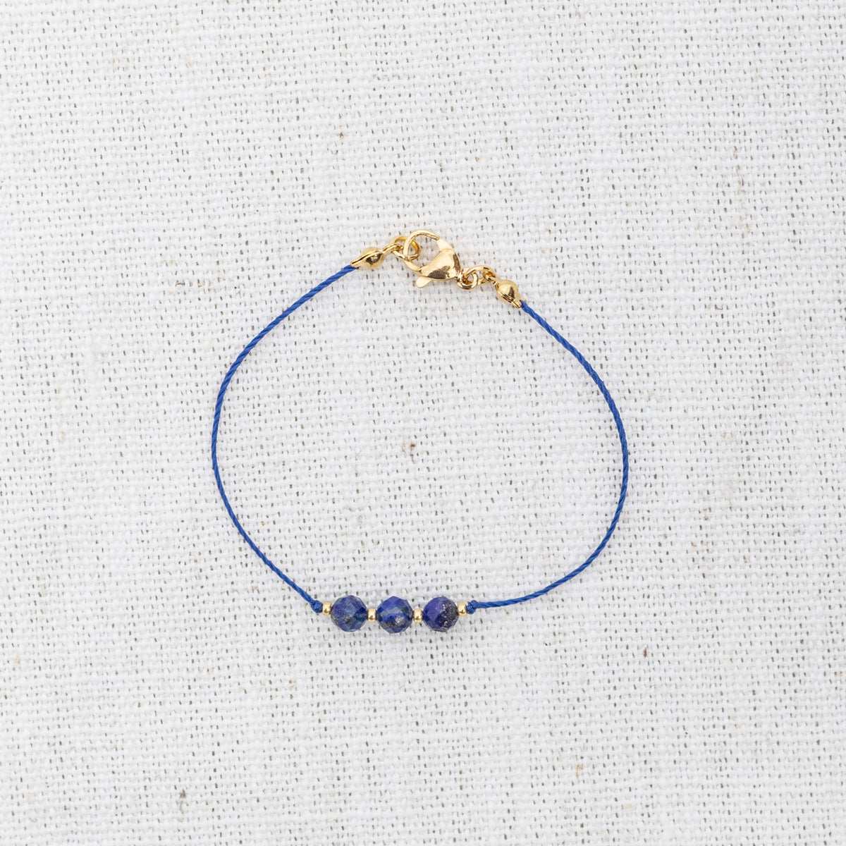 Intuitive Charms Amphitrite's Mist Lapis Lazuli Braided natural Crystal beaded custom Bracelet in gold and deep blue cord and clasp on a light fabric background