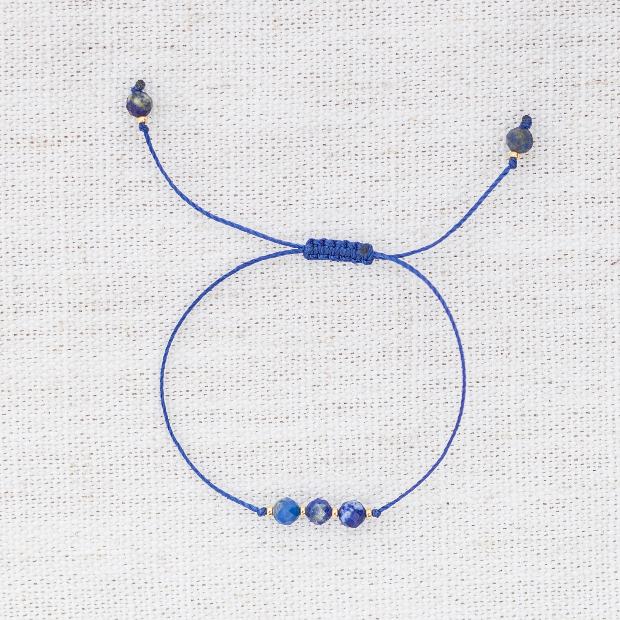 Intuitive Charms Amphitrite's Mist Lapis Lazuli Braided natural Crystal beaded custom Bracelet in gold and deep blue cord on a light fabric background