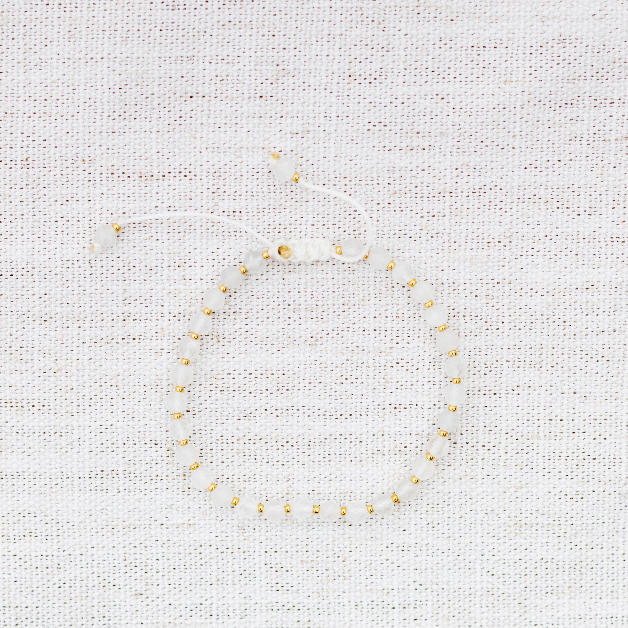 Intuitive Charms Selene's Glow Moonstone Braided natural Crystal beaded custom Bracelet in gold and white cord on a light fabric background