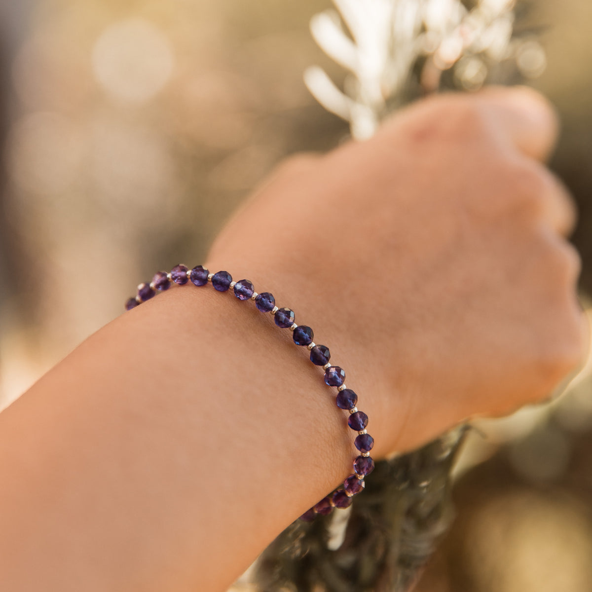 A close-up of a person's wrist wearing a beaded bracelet made of faceted dark purple amethyst gemstone beads, set against a soft, blurred natural background.