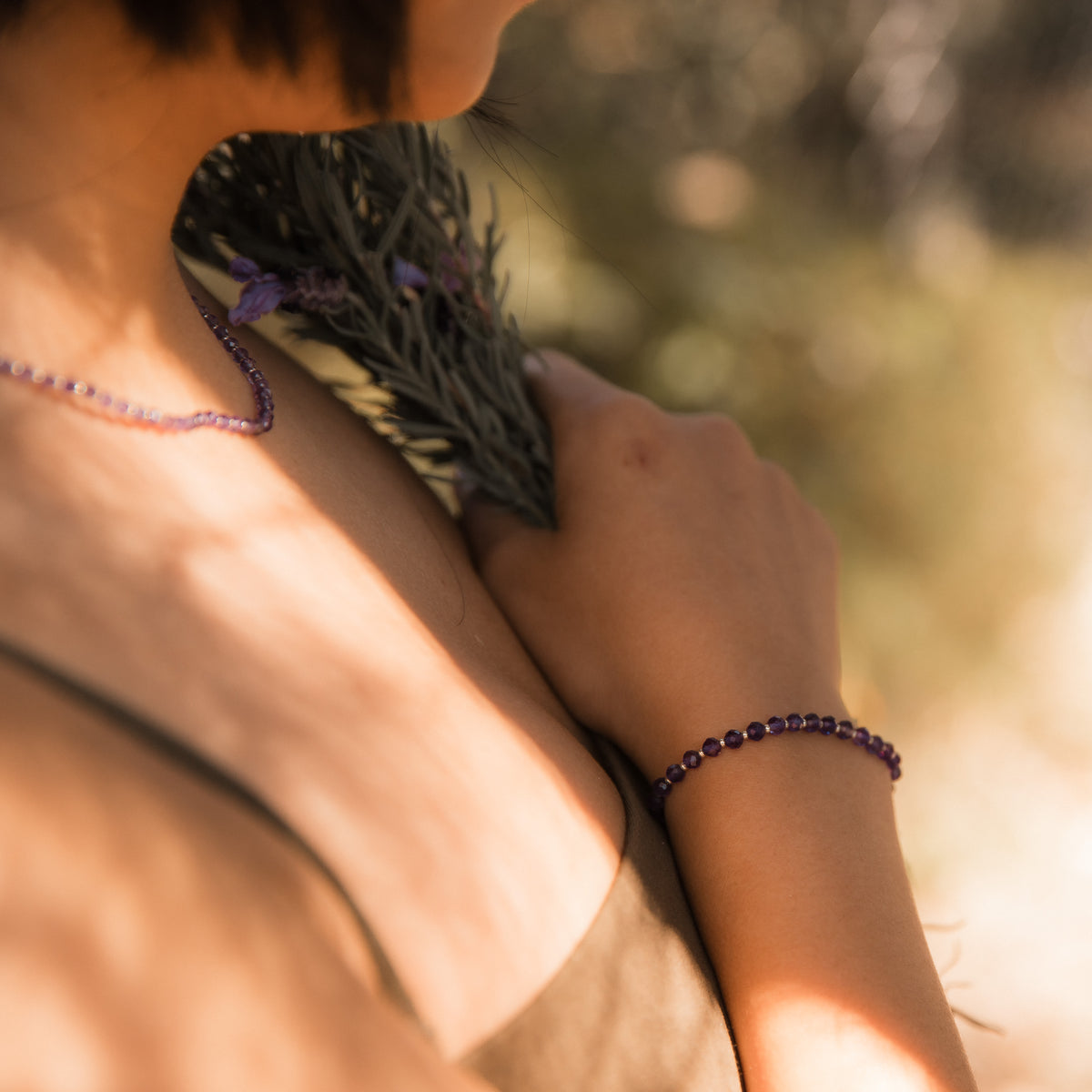 A close-up lifestyle shot of a person outdoors wearing a dark purple amethyst faceted beaded bracelet on their wrist and a matching necklace on their neck, holding a sprig of lavender. Made by intuitive charms.