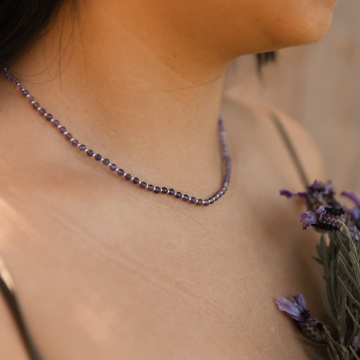 A close-up lifestyle shot of a person wearing a delicate beaded necklace made of small purple amethyst faceted beads, with lavender flowers resting on their shoulder against a warm, blurred background. Made by intuitive charms.