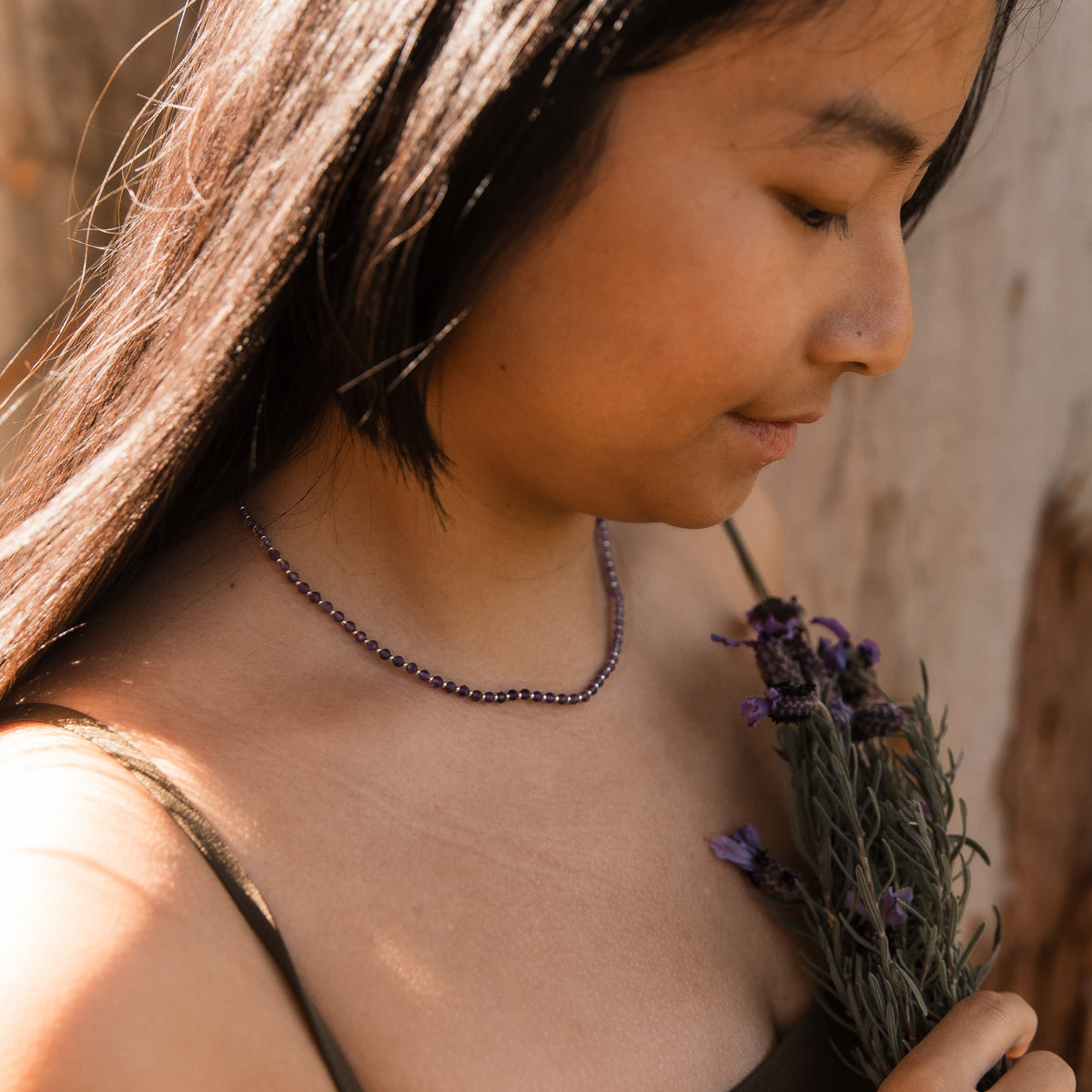 A close-up lifestyle shot of a woman wearing a delicate beaded necklace made of dark purple amethyst faceted beads. She is holding a small bunch of dried lavender flowers against a warm, rustic background. Made by intuitive charms.